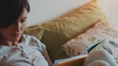 Genç kahverengi saçlı kadın yatak odasında uzanıyor ve kitap okuyor. Kızım oyuncaklarla oynuyor. Yeni tablet kullanan bir kız. Yakın plan..