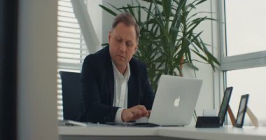 NEW YORK - 5 Nisan 2017: Modern beyaz ofiste masa başında oturan ve dizüstü bilgisayara bakan iş adamı. Pahalı takım elbiseli yakışıklı adam ayakta duruyor..