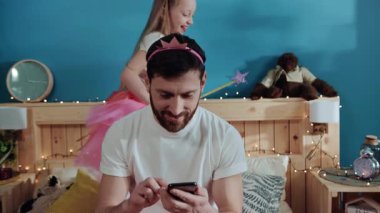 Yatakta otururken akıllı telefon kullanan sosyal bir adam. Prenses elbisesi içinde babasının etrafında dans eden mutlu küçük kız dikkatini vermiyor. Ev partisi kutlaması.