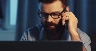 Gözlüklü bir adam laptopunun başında oturuyor, karanlıkta çalışırken bir telefon görüşmesi yapıyor.