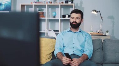 Bir adam rahat rahat rahat televizyon ekranına odaklanmış, elinde uzaktan kumandayla kanepede oturuyor.