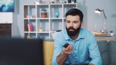 Bir adam kanepede rahatça dinlenirken elinde uzaktan kumandayla en sevdiği televizyon programının tadını çıkarıyor.