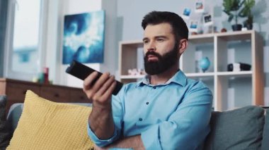 Düşünceli bir adam elinde uzaktan kumandayla rahat bir şekilde koltuğunda oturuyor ve evinde rahatlatıcı bir anın tadını çıkarıyor.