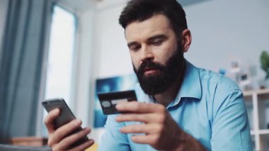 Bir adam internet üzerinden alışveriş yapmak için kredi kartı tutarken akıllı telefonunu anlaşmalara göz atmak için kullanıyor.