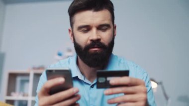 Odaklanmış bir adam ev ortamında kredi kartıyla online alışveriş yapmak için akıllı telefonunu kullanıyor.