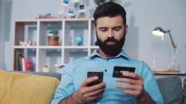 Odaklanmış bir adam evde online alışveriş yaparken akıllı telefonunu ve kredi kartını dikkatlice inceler.
