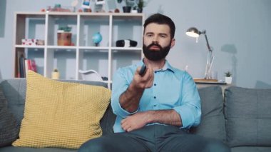 Mavi gömlek giyen neşeli sakallı bir adam TV izlerken rahat bir kanepede dinlenmenin tadını çıkarıyor.
