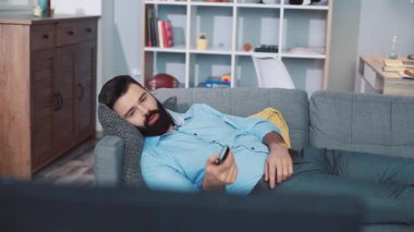 Bir adam rahat bir kanepede rahatlar, akıllı telefonuyla rahat yaşam alanında meşgul olur, keyfine bakar.