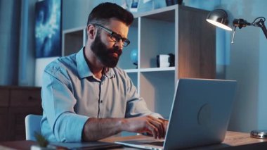 Gözlüklü bir adam gece geç saatlere kadar şık ve modern bir ofis ortamında dizüstü bilgisayarla çalışıyor.