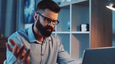 Ciddi, odaklanmış, gözlüklü profesyonel bir adam dikkatle dizüstü bilgisayarıyla loş bir ofiste çalışıyor.