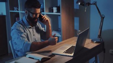 Gece geç saatlerde dizüstü bilgisayarı üzerinde çalışan ve aynı zamanda rahat bir çalışma ortamında telefonla konuşan bir adam.