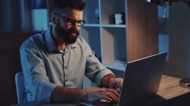 Gecenin sessiz saatlerinde loş bir odada oturan bir adam yoğun bir şekilde dizüstü bilgisayarı üzerinde çalışıyor.