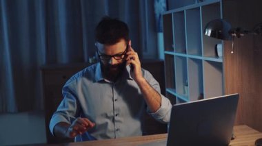 Kendini işine adamış bir adam gece geç saatlere kadar masasında çalışıyor. Notlarını düşünürken bilgisayarını kullanıyor.