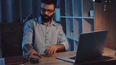 Bir adam, gece saatlerinde şık ve güzel aydınlatılmış bir ofiste özenle dizüstü bilgisayarı üzerinde çalışıyor.
