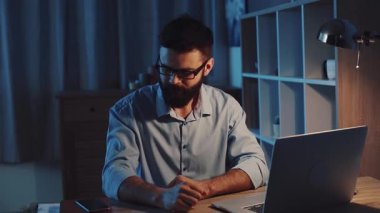 Mavi gömlekli bir adam masada oturuyor, kulaklık takıyor ve dizüstü bilgisayarına odaklanıyor.