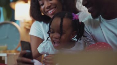 Çocuklu harika bir Afrikalı aile evdeki kanepede dinlenirken eğlenceli videolar izliyor. Anne baba ve kızı birlikte güzel vakit geçiriyorlar. Akıllı telefon kullanarak gülüyorlar..