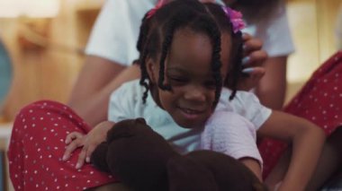 Annesinin dizlerinin üzerinde oturan oldukça yaramaz Afrikalı kız çocuğunun yakın çekimi. Genç anne kızının saçını düzeltmeye çalışıyor..