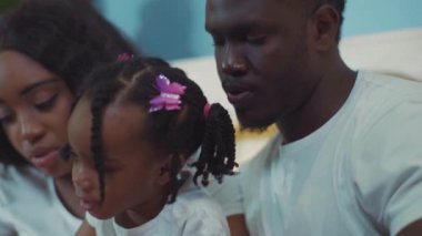 Odaklanmış güzel Afro-Amerikan anne ve babasının portresi ve evde yatakta birlikte kitap okuyan küçük bir kız..