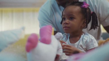 Örgü örmüş, babasıyla yatakta oturmuş etrafa bakınan Afro-Amerikan siyah bir kızın yakın plan portresi..