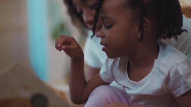 Saçı örgülü mutlu Afro-Amerikalı küçük bir kızın yakın çekimi. Annesiyle birlikte evdeki kanepede heyecanlı bir kitap okuyor..