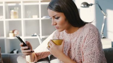 Kahverengi saçlı çekici genç bir kadın cep telefonunun ekranına bakıyor. Kız rahat giysiler rahat bir dairede kanepede oturuyor..