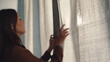 Genç, kahverengi saçlı, beyaz kadın sabah yatak odasında perdeleri açıyor. Pencereden bakan tatlı kız, gülümsüyor. Yeni güneşli gün.