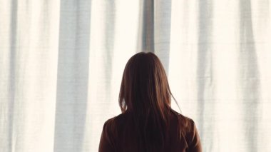 Gömlekli genç, kahverengi saçlı kadın geniş pencereye yaklaşıyor, sabah erkenden perdeleri açıyor. Pencereye bakan kız, yatak odasındaki şehir manzarasına hayran..