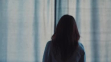 İş kıyafetleri içinde genç kahverengi saçlı kadın pencereye yaklaşıyor ve sabah perdeleri açıyor. Kız pencereden şehir manzarasına bakıyor. Kapalı.