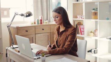 Genç, gözlüklü, modern tasarım ile aynı masada oturan, dizüstü bilgisayar kullanan çekici bir kadın. Şirin kız gülümsüyor. Kapalı.