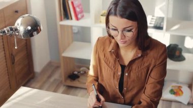 Gözlüklü ve gündelik elbiseli çekici genç bir kadın elinde lambayla masada oturuyor, dizüstü bilgisayarına bakıyor ve deftere bir şeyler yazıyor. Kapalı.