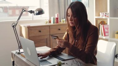 Elinde kredi kartı olan tatlı bir kız dizüstü bilgisayarın yanında oturuyor. Genç ve çekici bir kadın modern apartmandaki klavyede kredi kartı numarasını tanıtıyor..