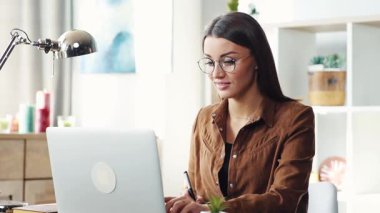 Gözlüklü ve kahverengi gömlekli çekici bir bayan öğrenci pazar günü masada oturup laptopta çalışıyor. Kapalı.