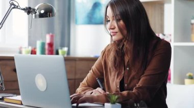 Masada oturan ve evdeki gümüş dizüstü bilgisayara bakan genç bir kadın. Klavyede yazan öğrenci.