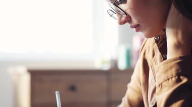 Yuvarlak gözlüklü, kahverengi gömlekli, ciddi düşünceli bir genç hanım masada oturuyor ve günlüğe not alıyor. İçeride. Yakın plan..