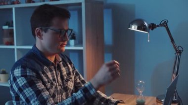 Gözlük takan odaklanmış bir adam, etrafını aydınlatma ve kum saati saran masasında özenle çalışıyor.