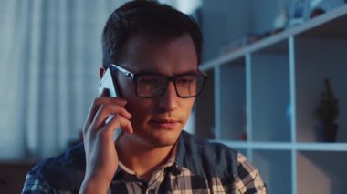 Gözlüklü genç bir adam, sıcak ve loş bir odada otururken önemli bir telefon görüşmesi yapıyor.