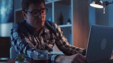 Yorgun bir birey masalarında dinleniyor, bir dizüstü bilgisayar ve içecekle çevrili, yumuşak ışıklı bir odada.