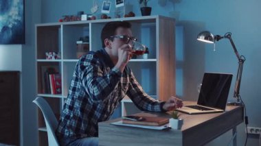Kendini işine adamış genç bir adam rahat ofisinde laptopuyla çalışırken içkisini yudumlamayı sever.