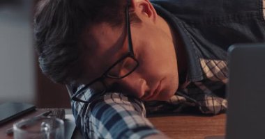 Yorgun bir genç adam yorgun başını dağınık masanın üzerinde dinleniyor, bir yığın kağıt ve içkiyle çevrili.