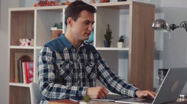 Şık ekose gömlekli odaklanmış genç bir adam modern ofis ortamında özenle dizüstü bilgisayarı üzerinde çalışıyor.