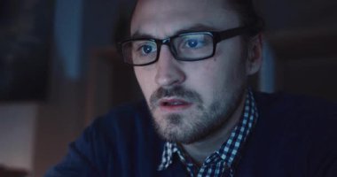 Gözlüklü bir adam yoğun konsantrasyon gösteren loş bir ortamda dikkatle dizüstü bilgisayarına odaklanır.