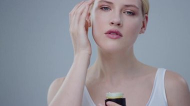 Nazik hareketlerle yeni krem sürmeye çalışan saf yüzlü, güzel, narin sarışın kızın yakın çekimi. Cilt bakımı için kozmetik ürünü kullanan model kızın portresi ve gri arka planda mutluluğun gülümsemesi..