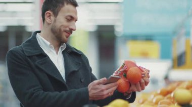 Markette elinde alışveriş arabasıyla dolaşan yakışıklı genç adam. Telefon üzerinden alışveriş listesine bakıyor. Meyve alıcısı, yiyecek dükkanı, market, perakende satış mağazası, yavaş çekim.