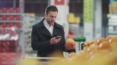 Telefondaki alışveriş listesine bakan alışveriş arabasıyla süpermarkette yürüyen gülümseyen genç bir adam. Meyve alıcısı, yiyecek dükkanı, market, perakende satış mağazası, yavaş çekim.