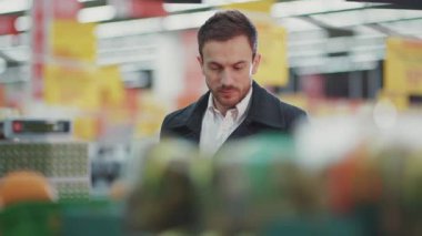 Markette elinde alışveriş arabasıyla yürüyen genç adam telefon ışığında alışveriş listesine bakıyor. Meyve alıcısı seçiyor.