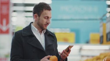 Markette elinde alışveriş arabasıyla gezen genç bir portre. Telefon ışığında alışveriş listesine bakıyor. Meyve alıcısı, yiyecek dükkânı, yetişkin, perakende satış mağazası, yavaş çekim.