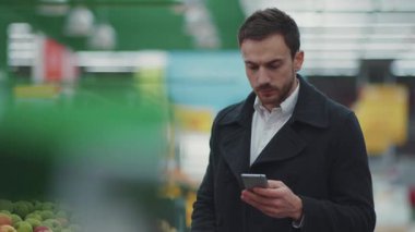 Süpermarkette elinde alışveriş arabasıyla gezen genç adam telefon ışığında alışveriş listesine bakıyor. Meyve alıcısı seçiyor.