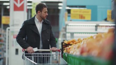 Süpermarkette elinde alışveriş arabasıyla dolaşan genç adam, telefondaki alışveriş listesine bakıyor. Meyve alıcısı, yiyecek dükkanı, alışveriş mağazası, yetişkin perakende satış mağazası, yavaş çekim.