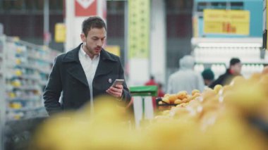 Gözlüklü ve gömlekli sakallı adam elinde alışveriş arabasıyla süpermarkette yürüyor. Akıllı telefondan alışveriş listesine bakıyor.