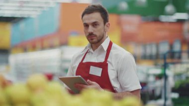 Satıcı, süpermarkette tablet bilgisayar kullanır. Kameraya bak. Sebzeler, sebzeler, sebzeler...